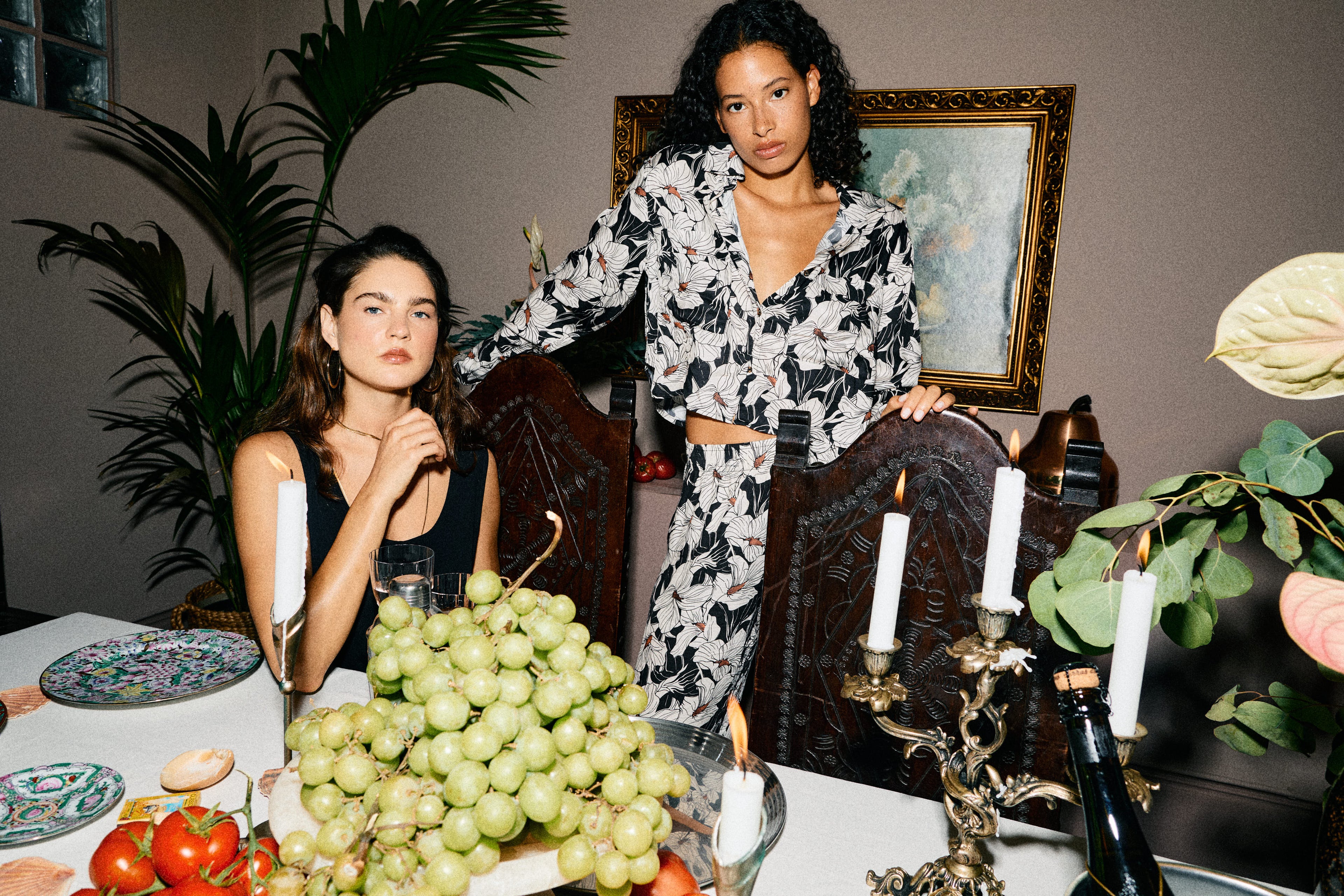 Two women sitting at a table with fruits and candles, with a painting in the background.