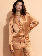 apricot-washed-satin- Woman wearing the Casa Lélé Marais blazer in apricot satin with matching trousers against a beige background