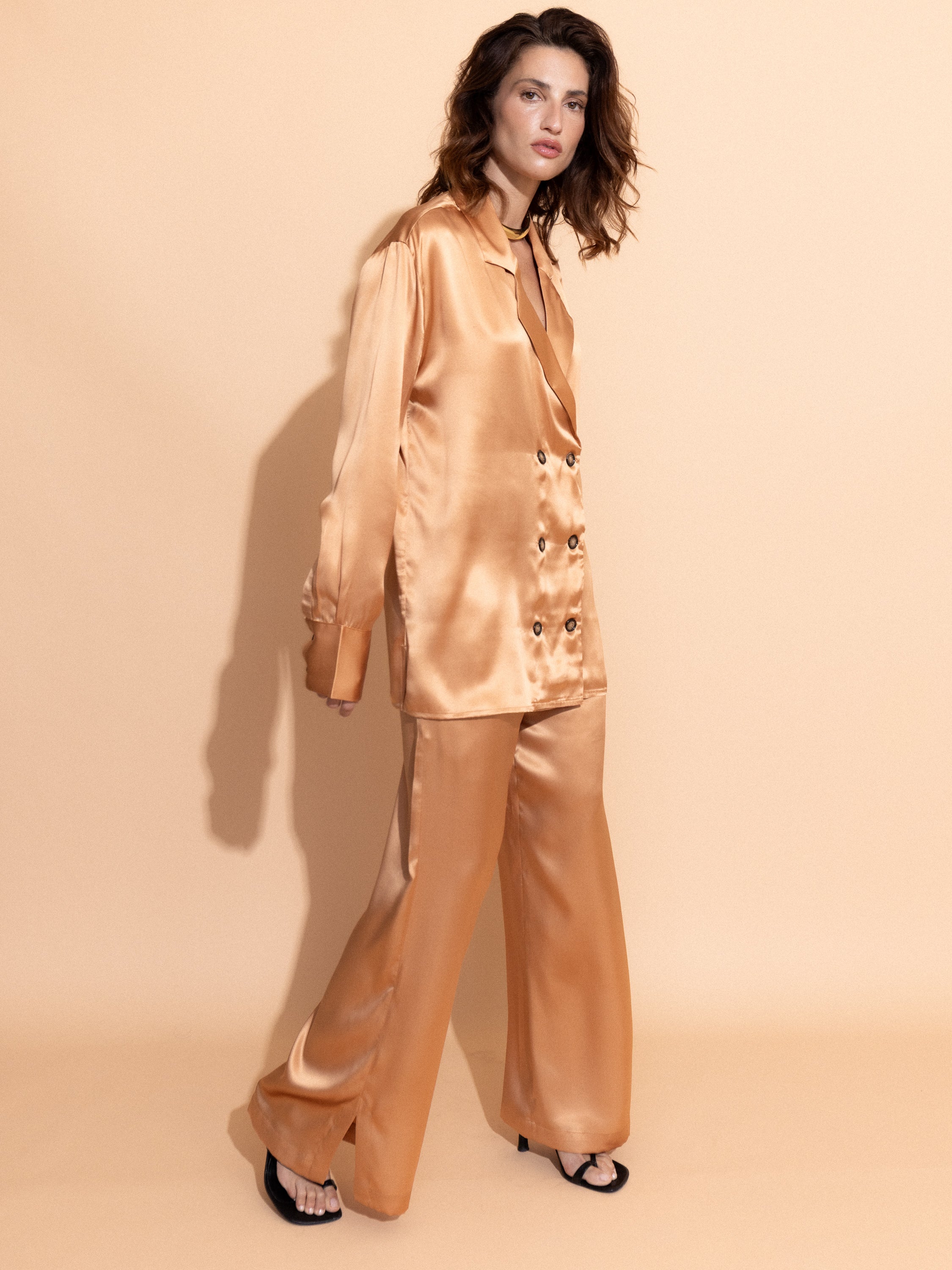 apricot-washed-satin - Woman wearing the Casa Lélé Marais blazer in apricot satin with matching trousers against a beige background
