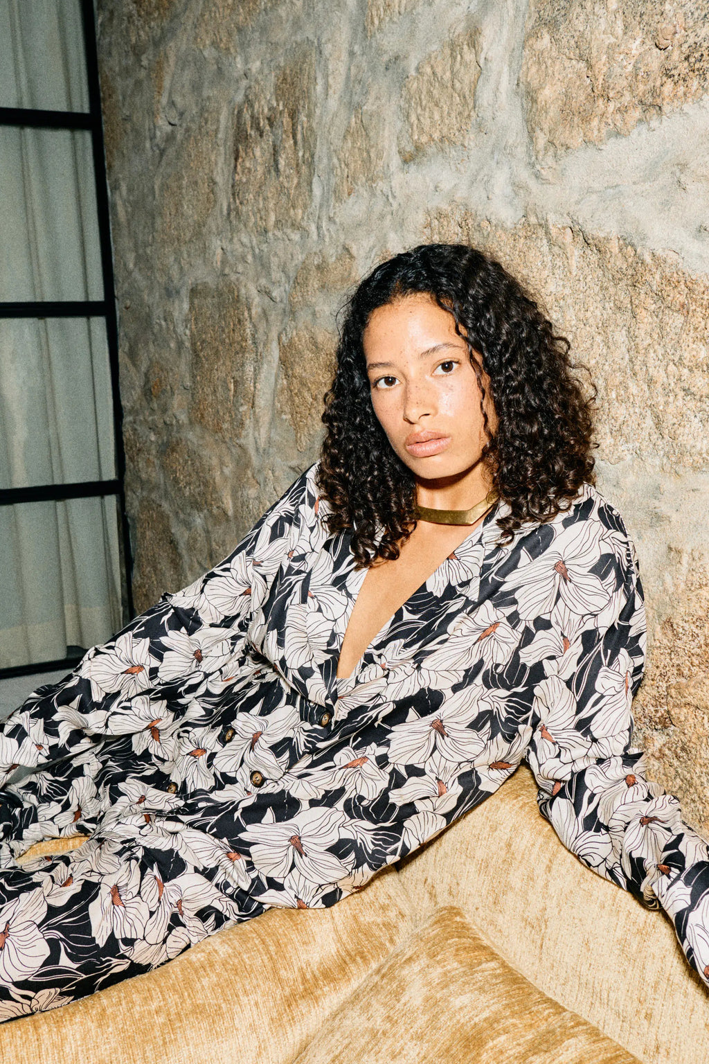 Woman in a floral two piece suit sitting on a couch against a stone wall.