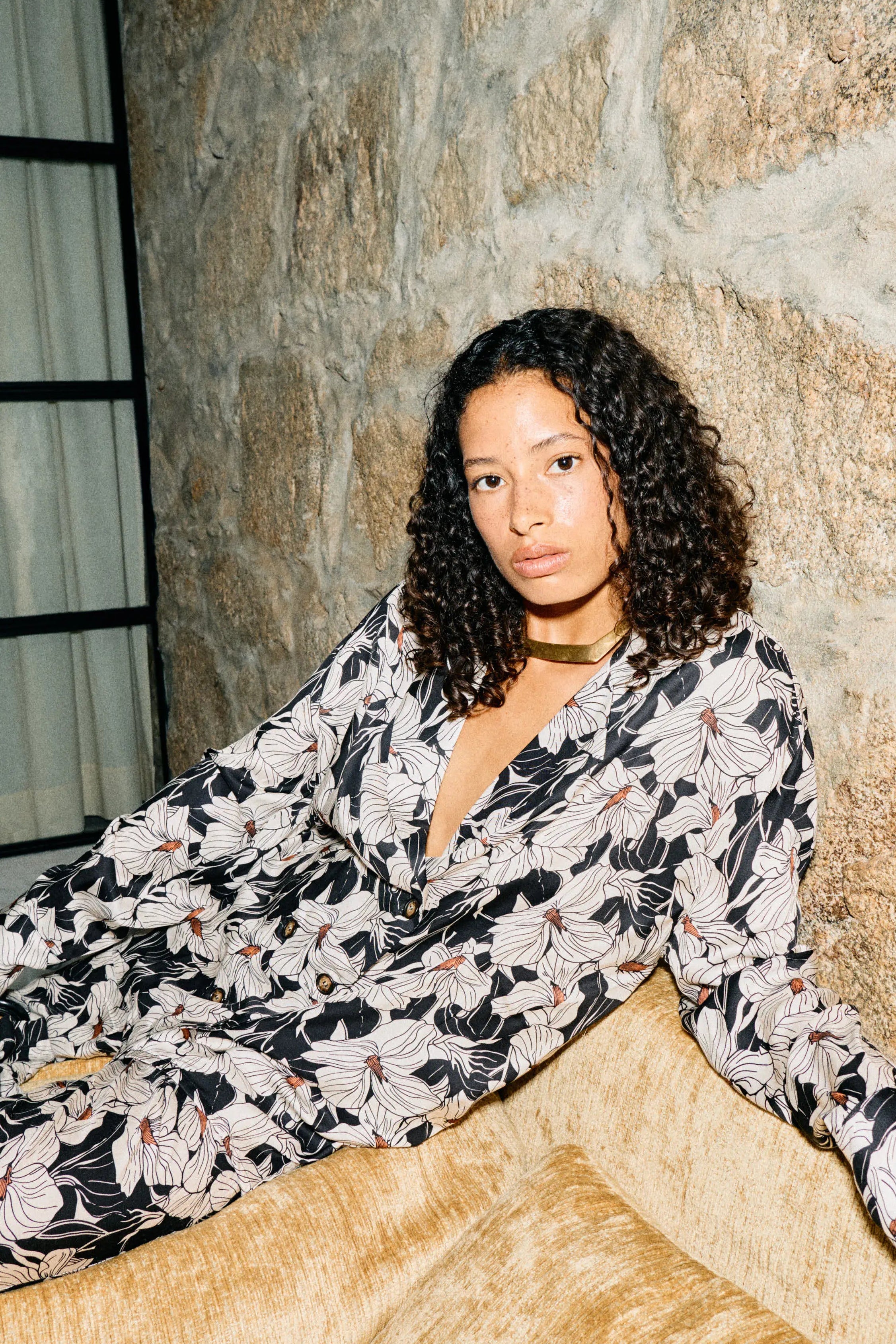Woman in a floral two piece suit sitting on a couch against a stone wall.