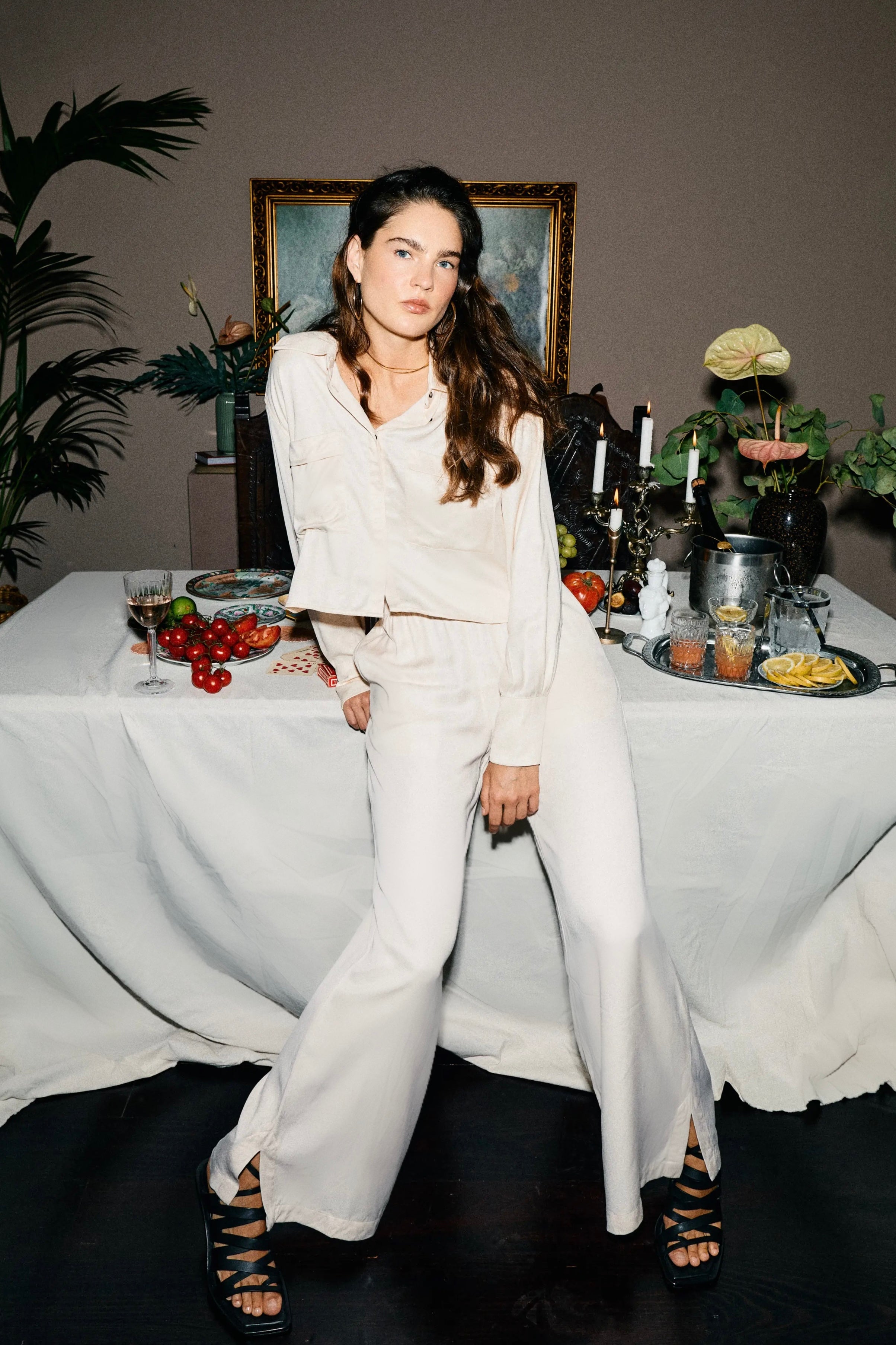 Woman in a white cream casa lélé matching cream loungewear set standing in front of a decorated table with plants and art in the background.