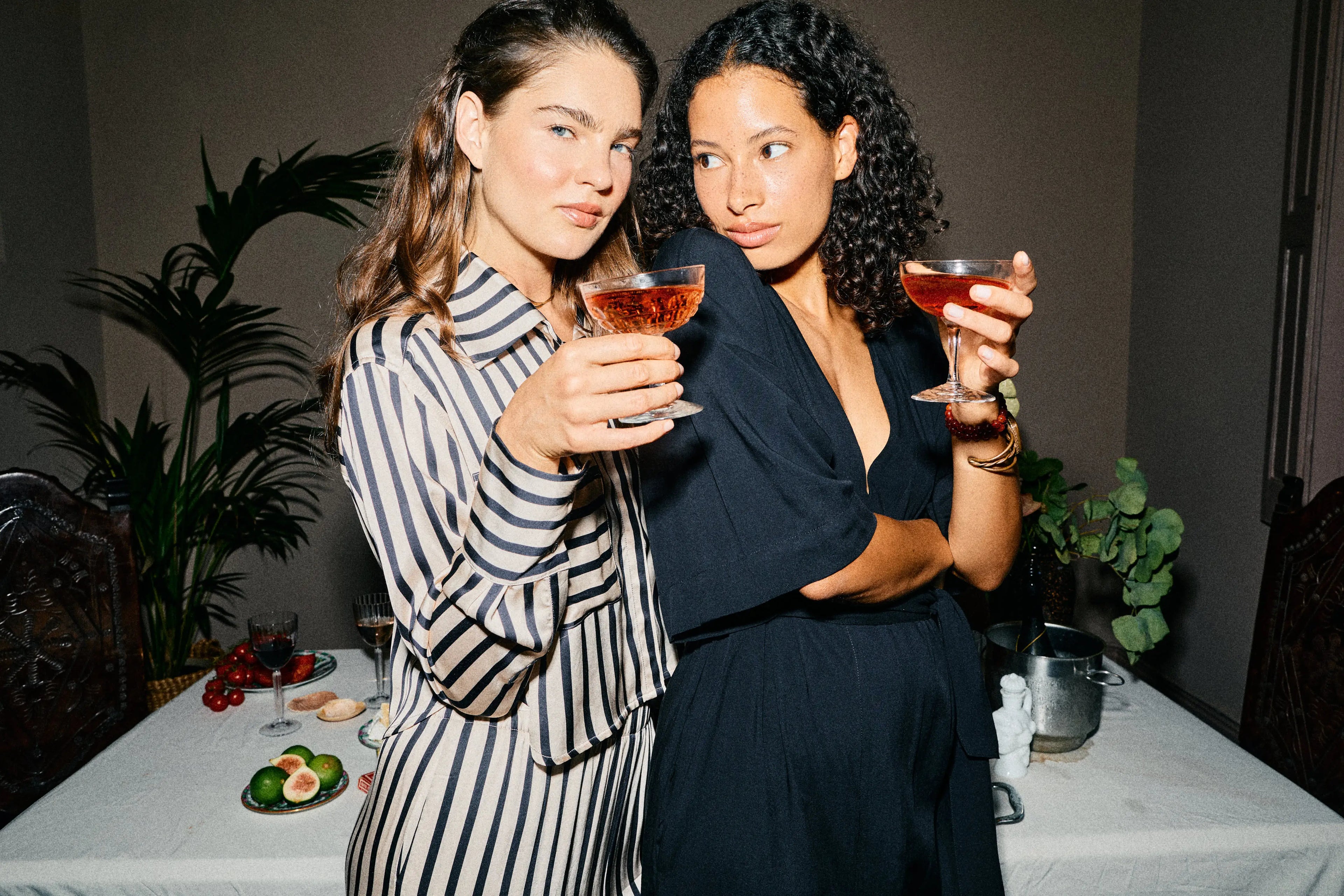 Two women at a dinner table holding drinks, wearing Casa Lélé Cleo Cropped Top and Lana Kaftan Dress with a plant and table setting in the background