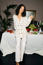 Woman in a white outfit standing by a table with food and drinks, holding a glass.
